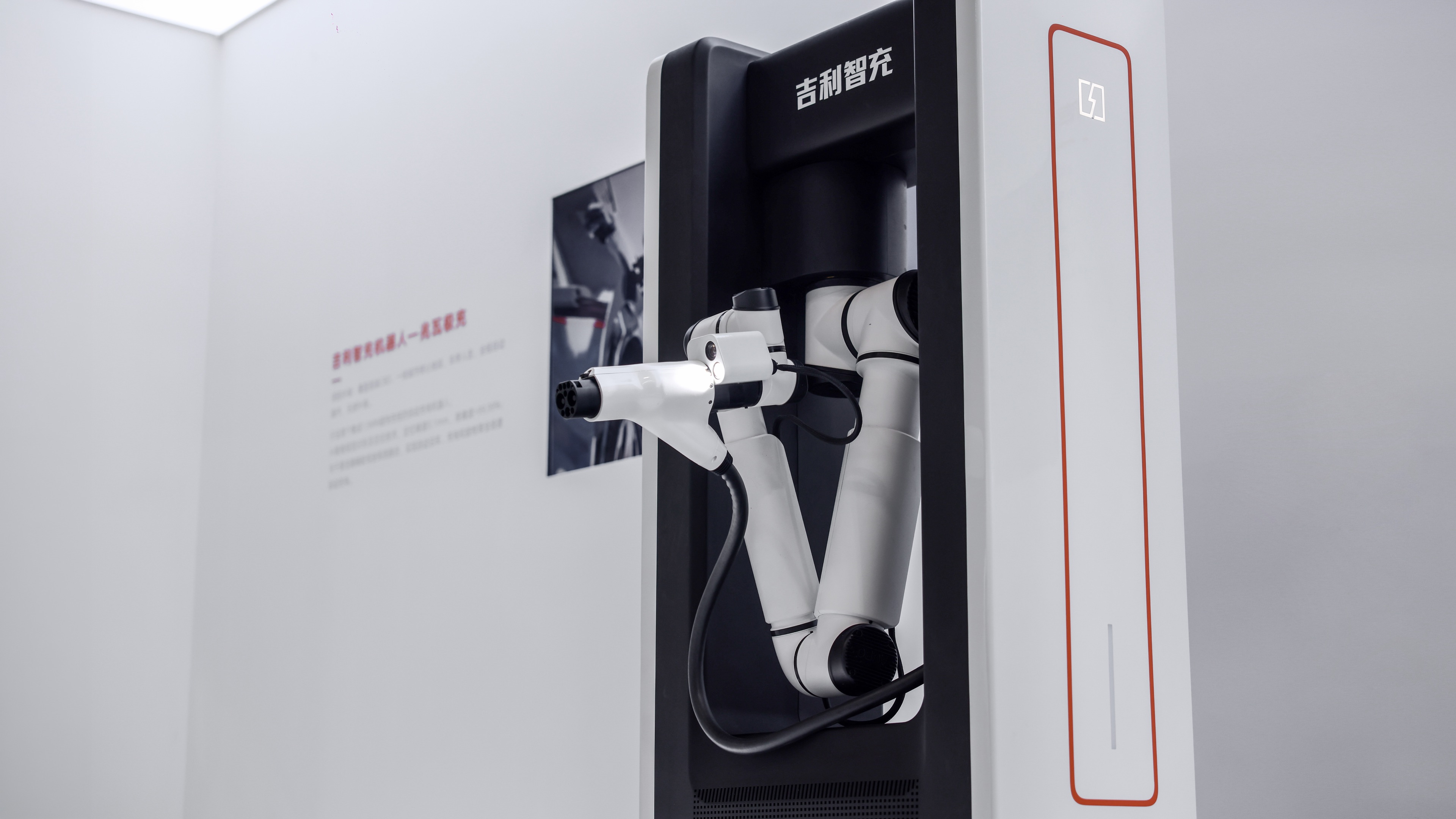 Geely C12 şarj ünitesi ve konektörü tutan beyaz robot kol, kapalı sergi alanında