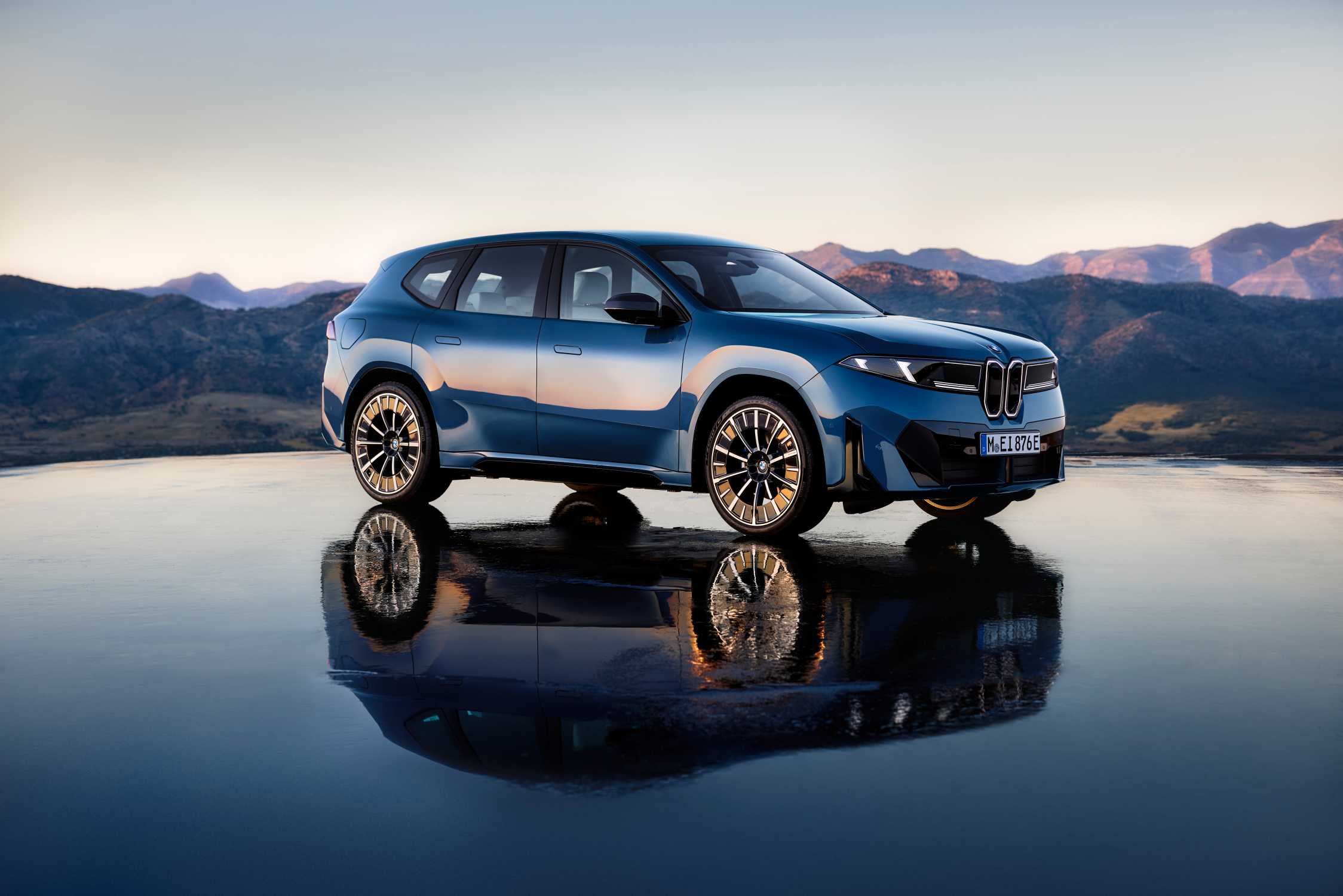 Mavi BMW iX3 50 xDrive'ın dağ manzarası önünde parlak zeminde üç çeyrek görünümü