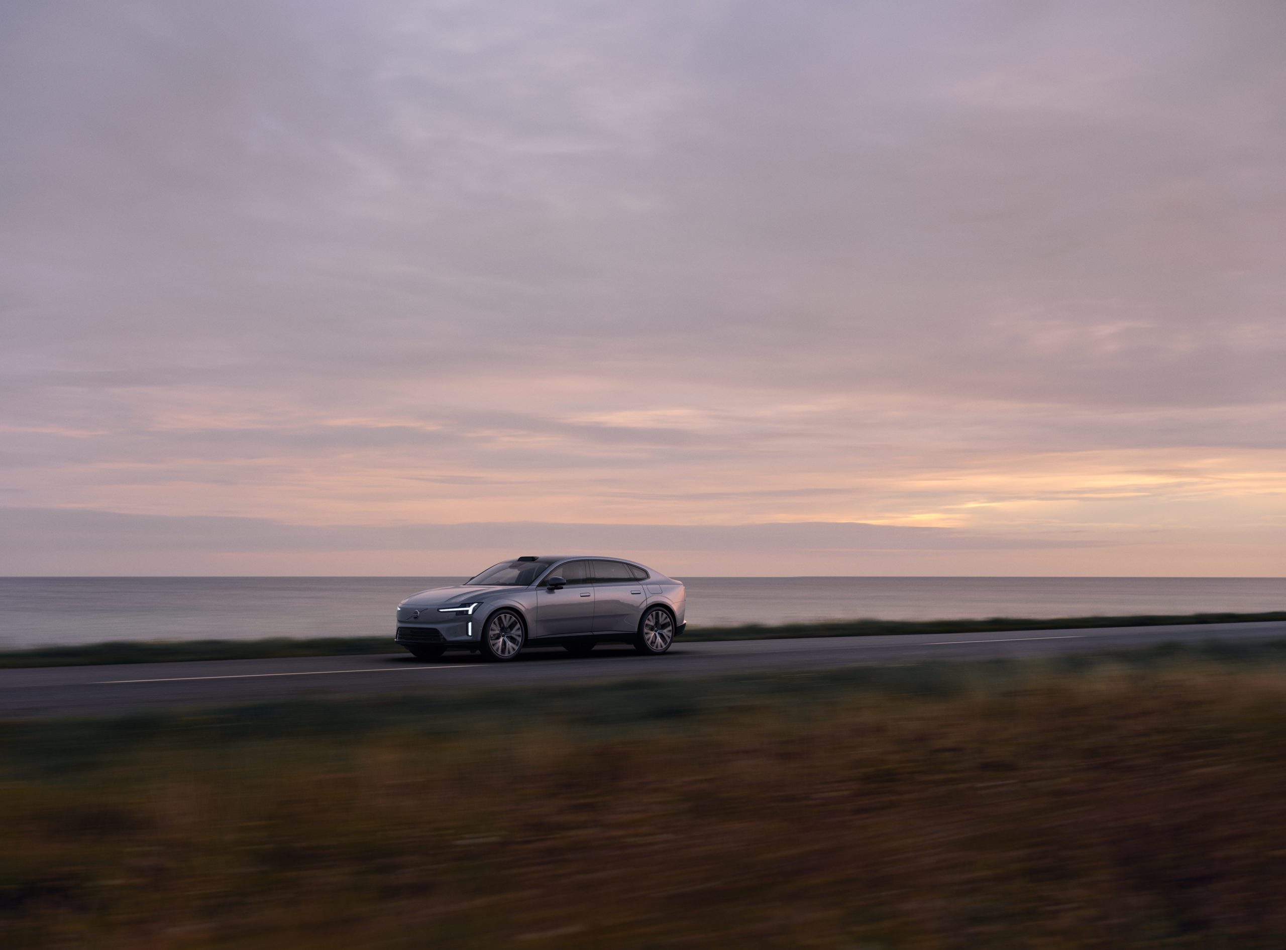 Gümüş gri Volvo ES90 alacakaranlıkta deniz kıyısındaki yoldan geçerken çekilmiş hareket fotoğrafı