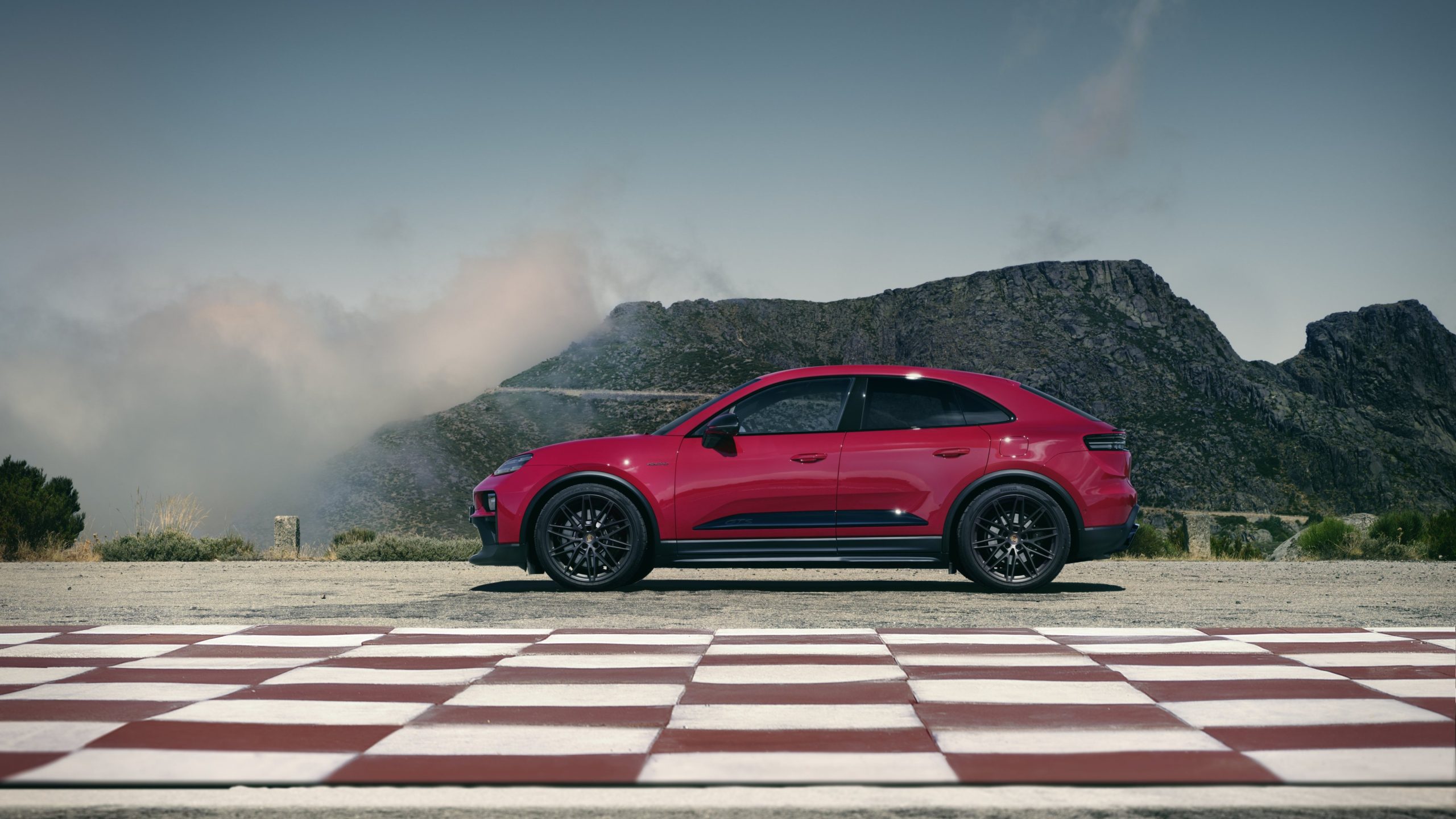 Kırmızı elektrikli Porsche Macan GTS damalı zemin üzerinde dağ manzarasına karşı yandan görünümde