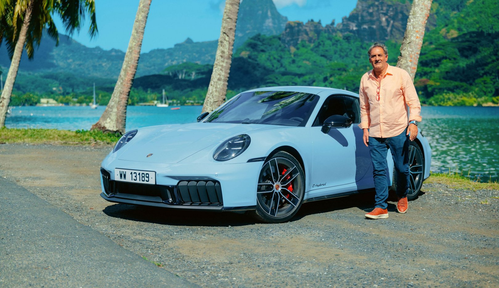 Açık mavi Porsche 911 Carrera GTS, palmiyelerin ve bir körfezin önünde duruyor; yanında turuncu gömlekli bir adam var