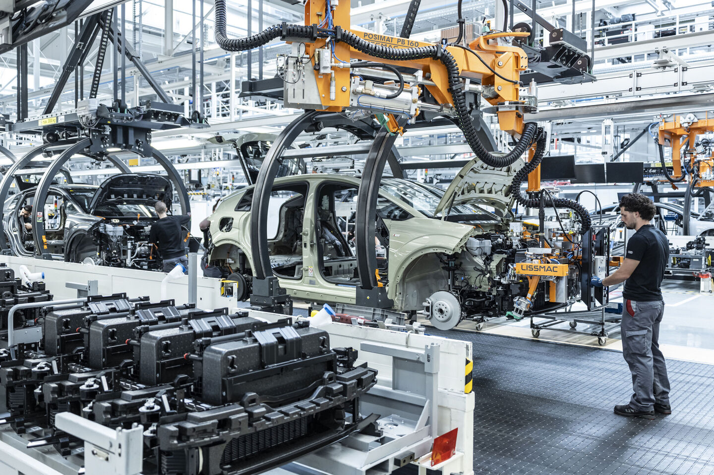 Audi A2 e-tron kaporta atölyesinde sarı ve turuncu robot kolları, kaporta parçalarını taşıyan yarı mamul bir gövdenin çevresinde çalışıyor