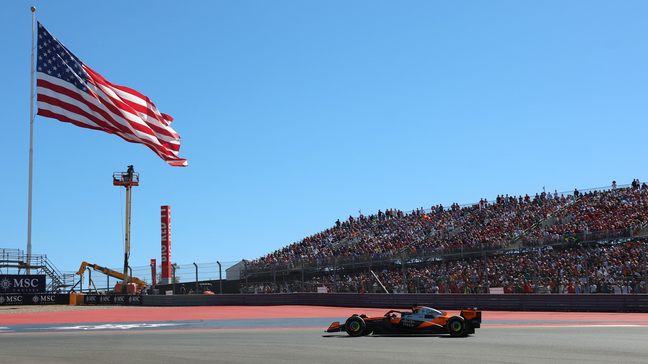 Austin’de Circuit of The Americas pistinde ABD bayrağı ve tribünlerin önünden geçen turuncu bir Formula 1 otomobili görülüyor