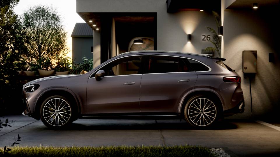 Gri renkli Mercedes-Benz elektrikli SUV akşam ışığında evin önünde yandan görülüyor ve duvar şarj cihazına bağlı