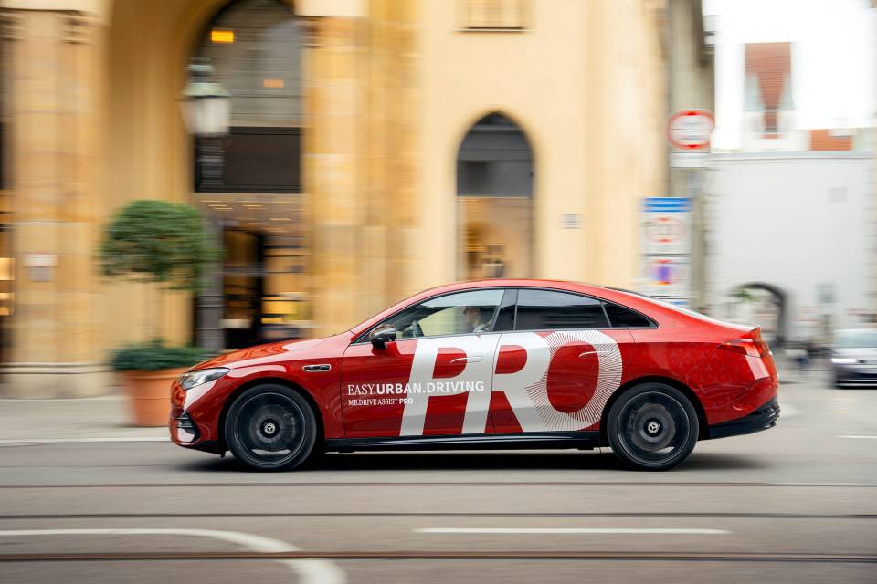 Kırmızı Mercedes-Benz, MB.Drive Assist Pro tanıtımında şehir içinde yan profilden hareket hâlinde görünüyor