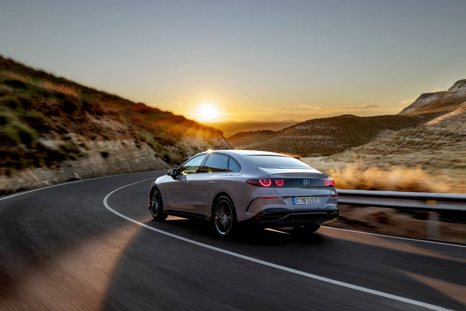 Elektrikli Mercedes C 400 4MATIC sedanın gün batımında kıvrımlı yolda arkadan üç çeyrek görünümü