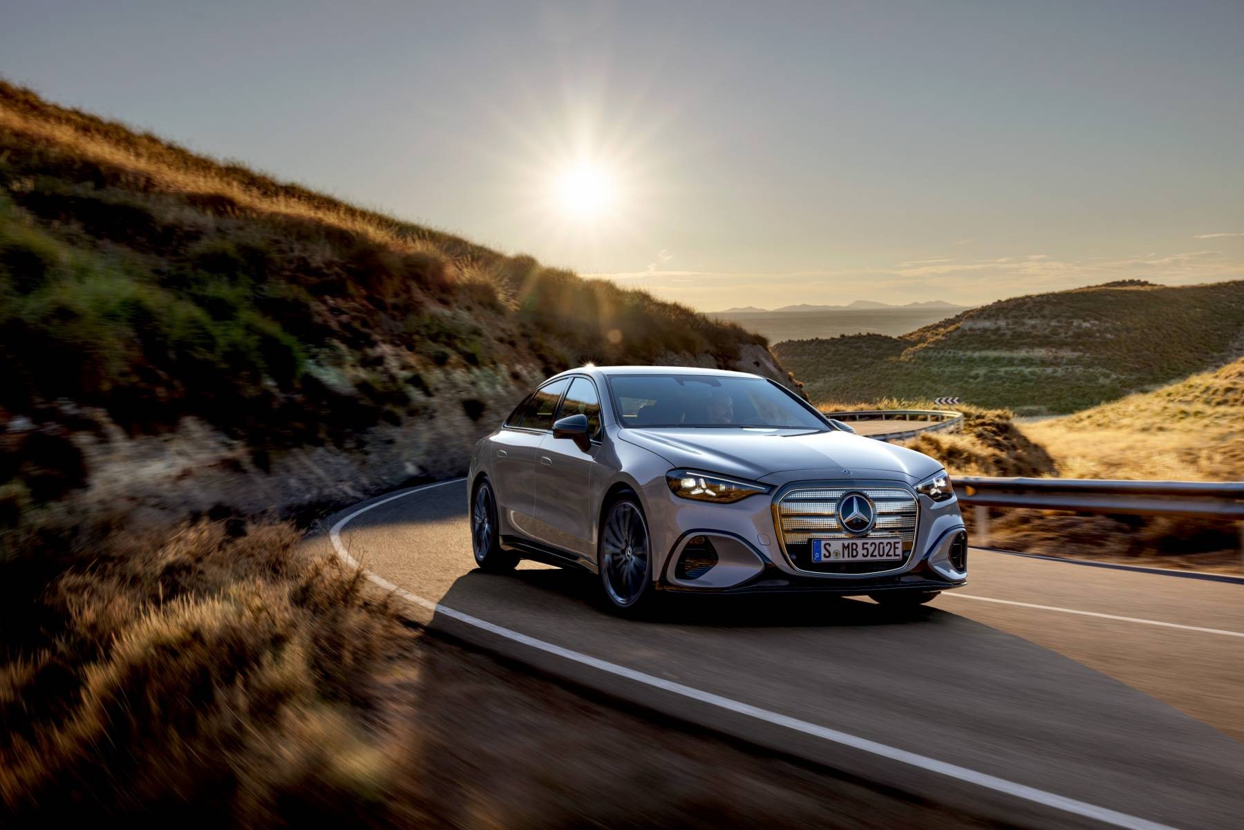 Gümüş rengi Mercedes C 400 4MATIC elektrikli sedanın güneşe doğru ilerlerken önden görünümü